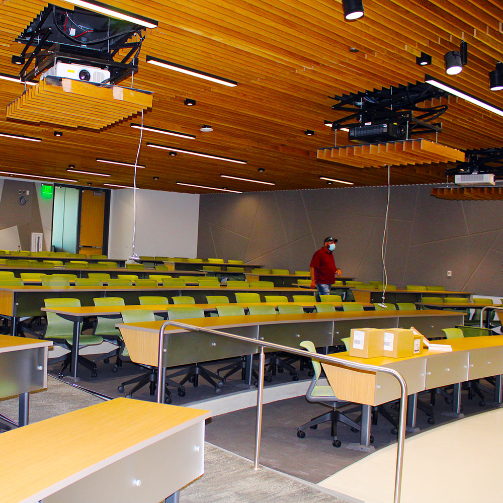 A worker in protective equipment walks through a lecture hall where wires and pieces of audio-video equipment hang from the ceiling.