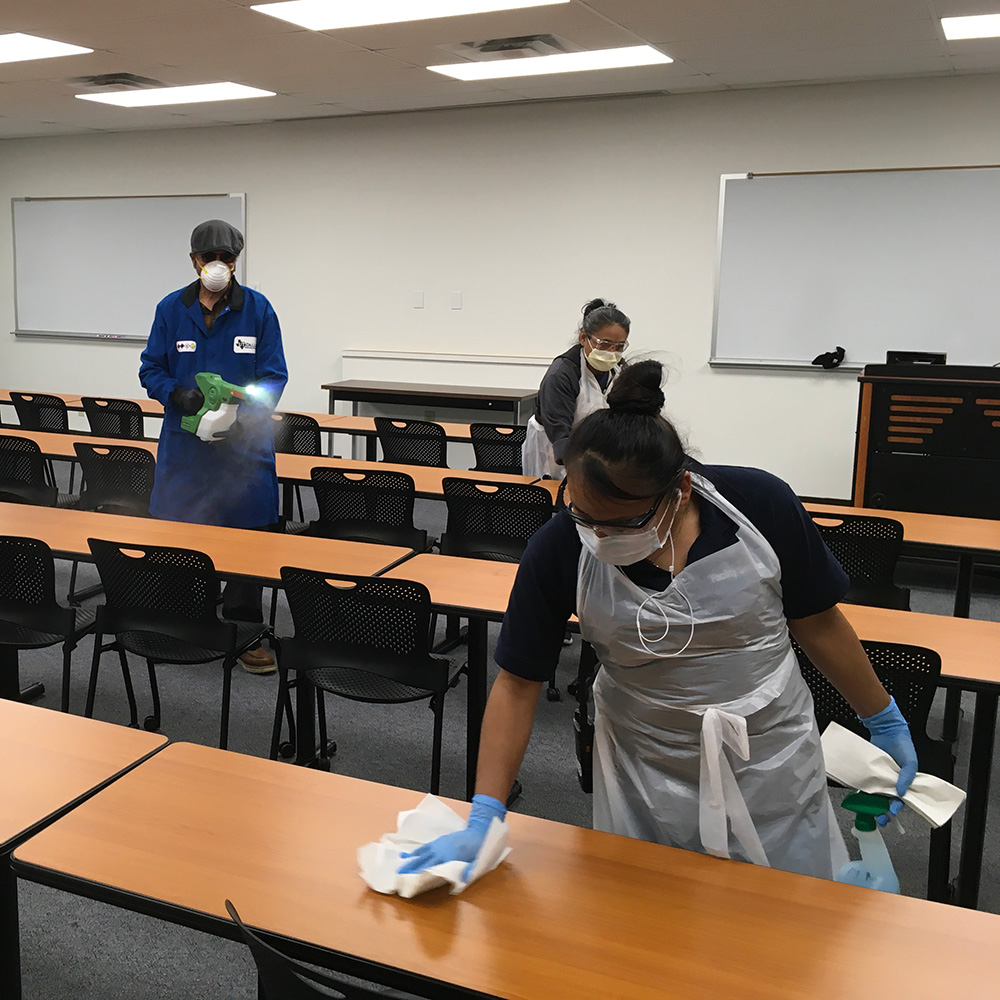 Custodial workers in personal protective equipment sanitize a classroom.