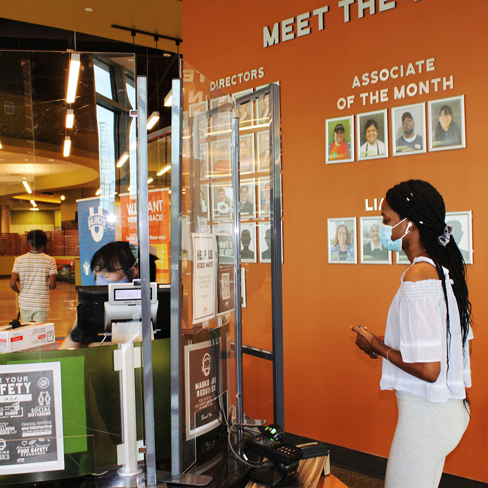 A student and a Dining Services worker at the hands-free counter in Dining Hall West, both wearing masks and separated by plexiglass.