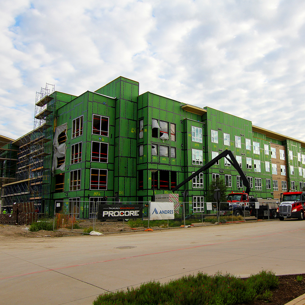 A multi-story building covered in scafolding as a layer of green panels are being applied to its exterior.