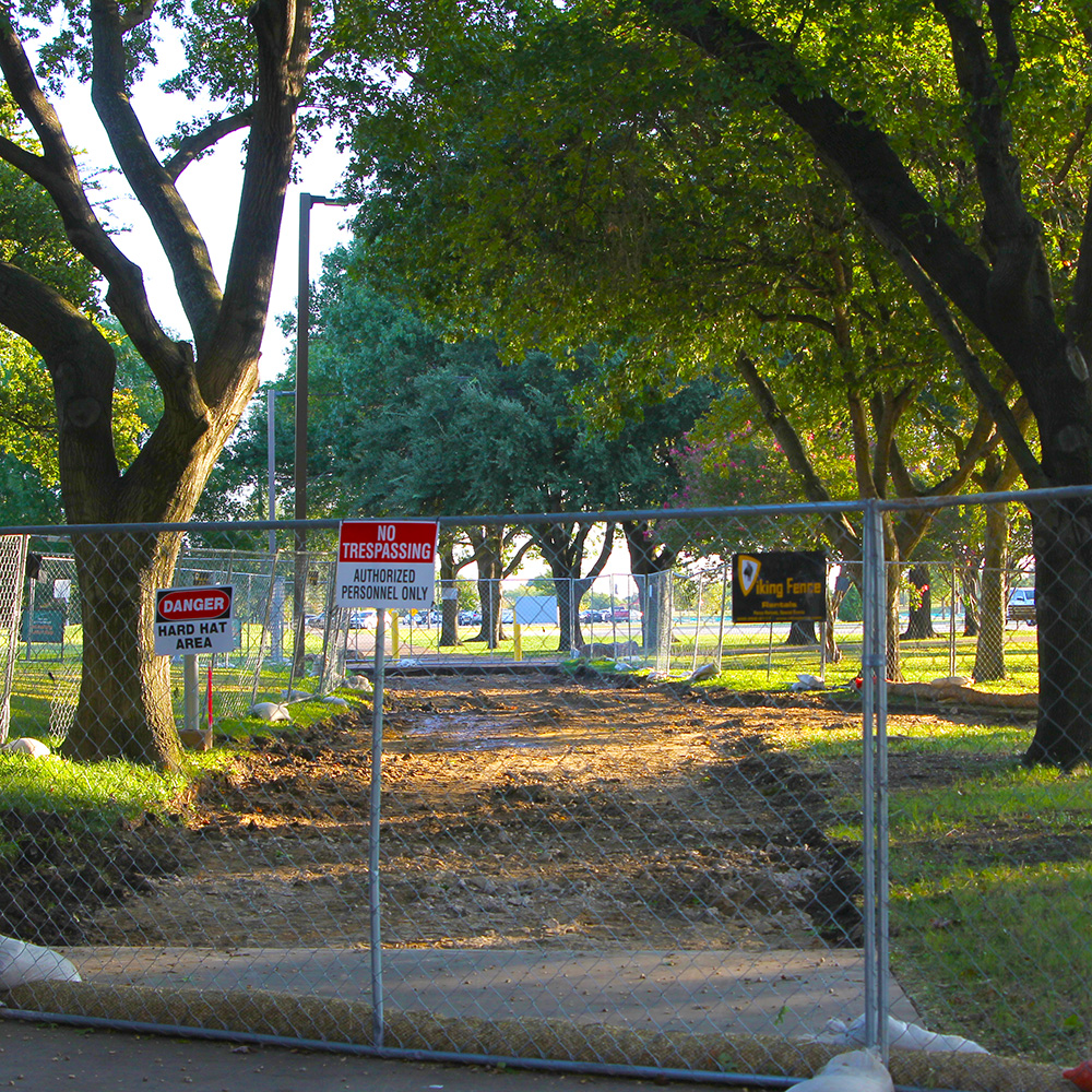 An angular dirt path, cut into the sod between rows of trees, all of which is surrounded by fences.