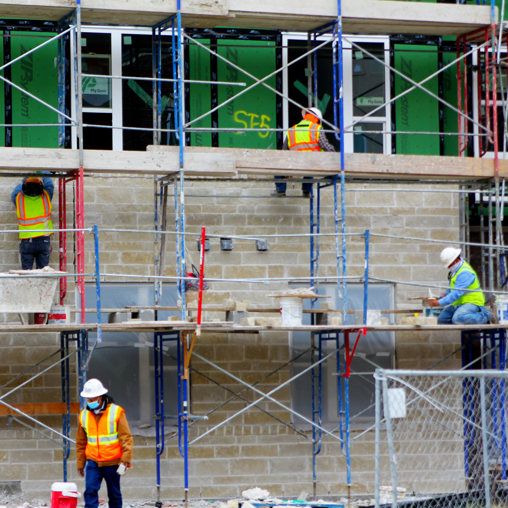 Work continues on the latest phase of Northside at UT Dallas. Four workers on scafolding work on the stone exterior of a building.