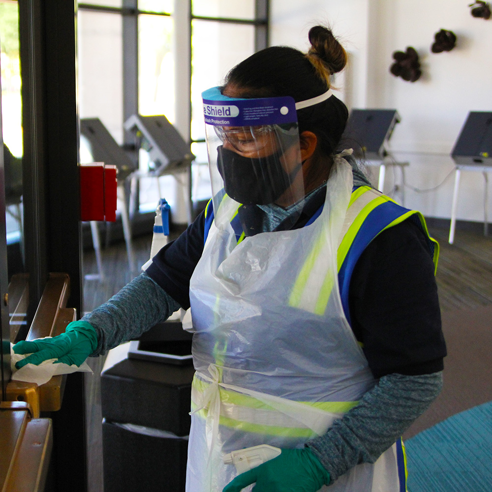 A member of the custodial crew in full personal protective equipment sanitizes a door with a spray bottle and wipes.