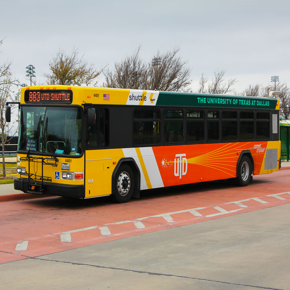 A long blocky bus, coverd in a mix of the yellow-and-white DART design and the orange-and-green UT Dallas design.