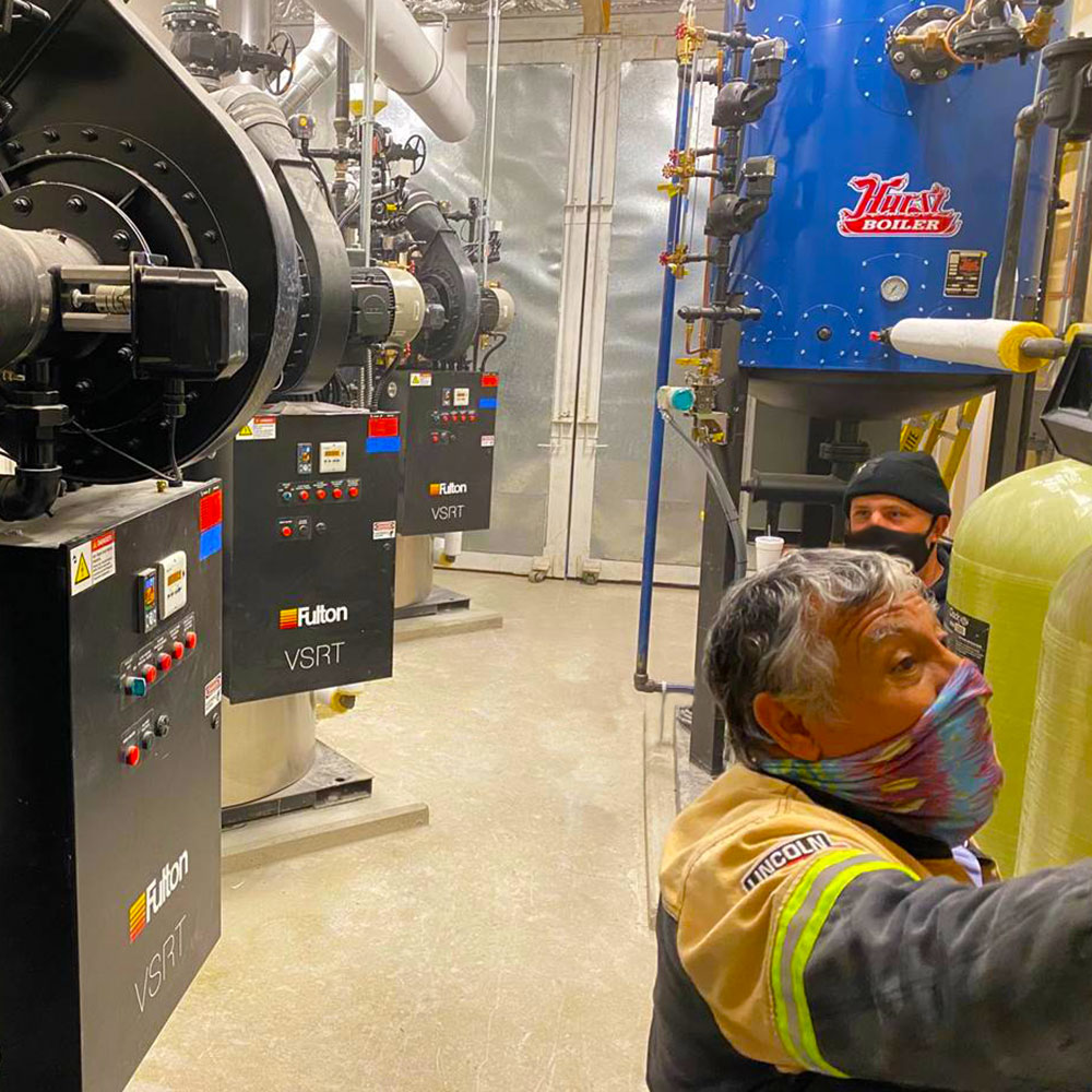 A pair of workers in Personal Protective Equipment between boilers and pipes.