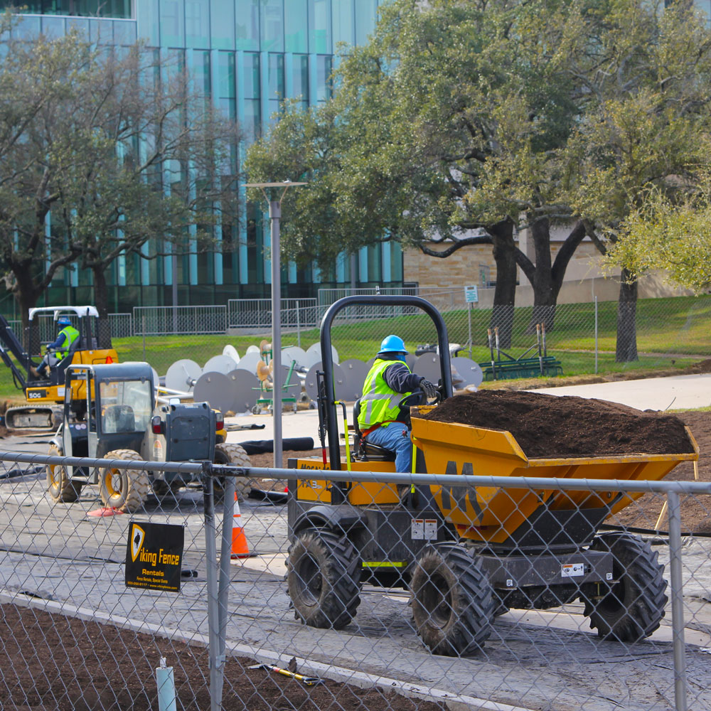 Construction equipment moves dirt over what used to be a street between academic buildings, while streetlamps rest on their sides at the edge of the construction zone.