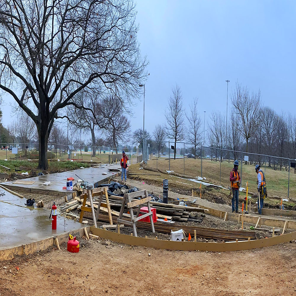 Construction workers check on newly poured concrete sidewalks and on a series of wooden molds laid on the ground in preparation to recieve more concrete.