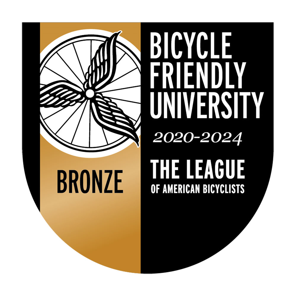 A logo featuring three feathered wings radiating out from the center of a spoked bicycle wheel, with words reading: Bicycle Friendly University, 2020-2024, The League of American Bicyclists, Bronze.