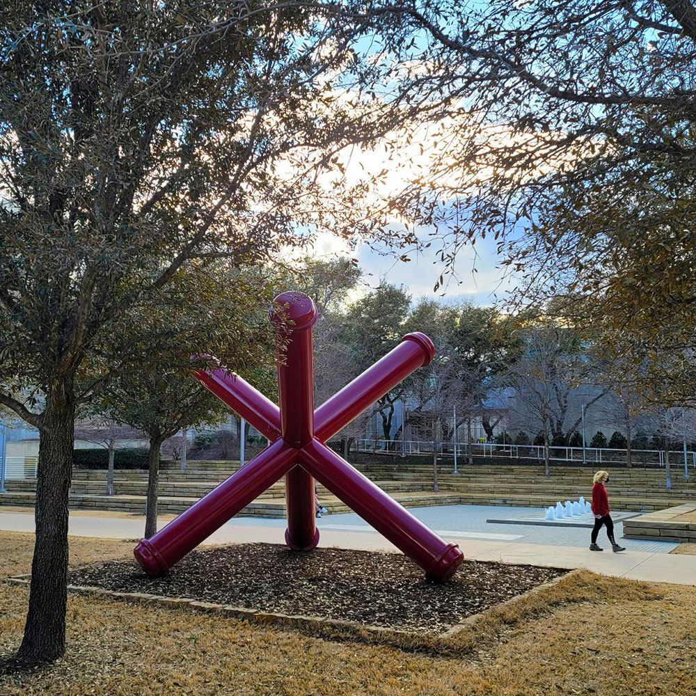 An abstract red metal sculpture, surrounded by trees, in front of a group of fountains.