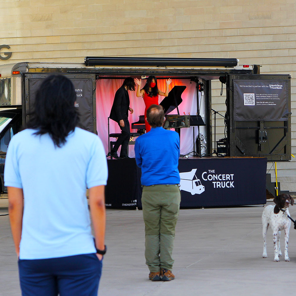 A small black cargo truck, with one side completely opened by a pair of doors that stretch its full length, reveals a small stage inside complete with a grand piano, gauzy background, and a pair of pianists in protective masks. In front of the truck a group of people, each spread far apart from one another, applaud the performance.