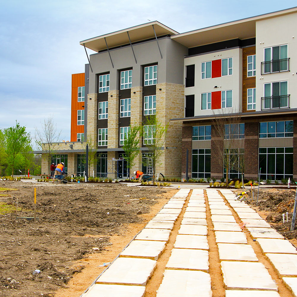 A contemporary four-story apartment building finished in multiple materials colored brown, cream, grey, and orange; before which a limestone brick path leads forward into a grass-covered park.