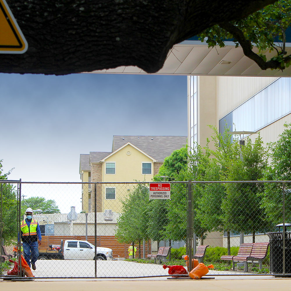 Seen from under the bough of a large tree and behind a chain-link fence, a worker in safety gear walks between two rows of facing benches and trees.