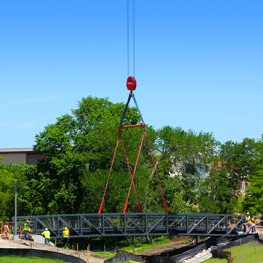 A curved black metal bridge is lowered by wire over the two sides of a creek while construction workers pull on both ends to guide it into its final position.