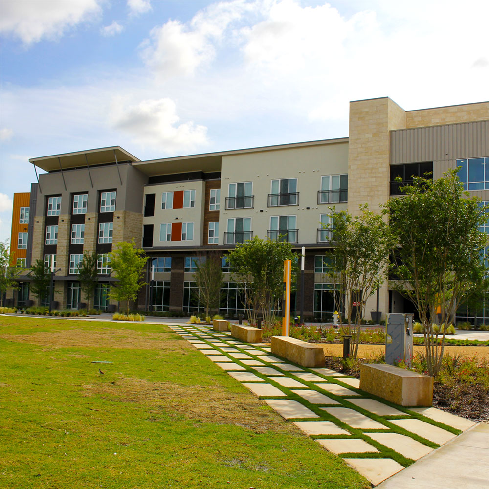 A contemporary four-story apartment building finished in multiple materials colored brown, cream, grey, and orange; before which a limestone brick path leads forward into a grass-covered park.