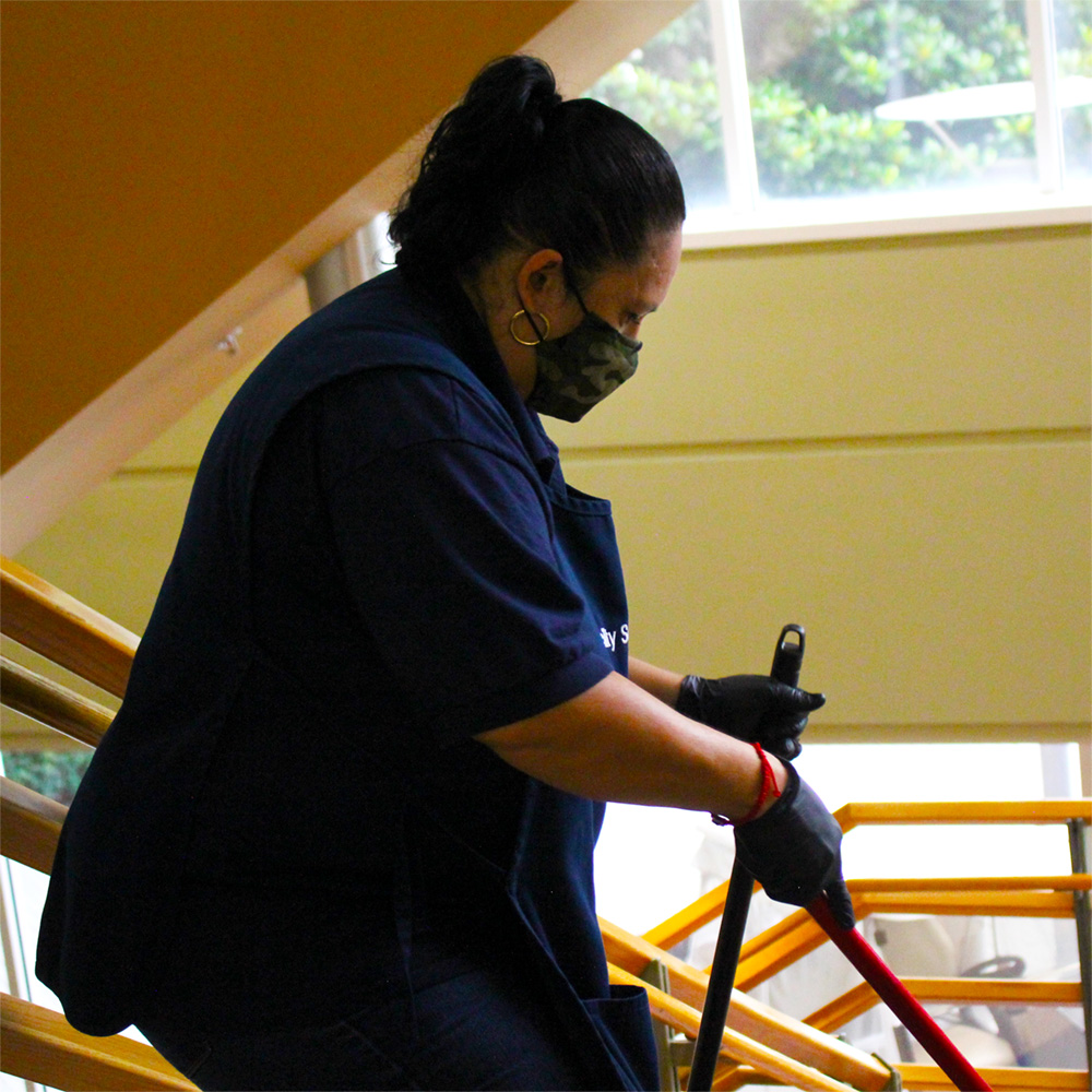 A woman in a custodian apron, mask and gloves sweeps a stairwell using a long-handled dustpan and broom.