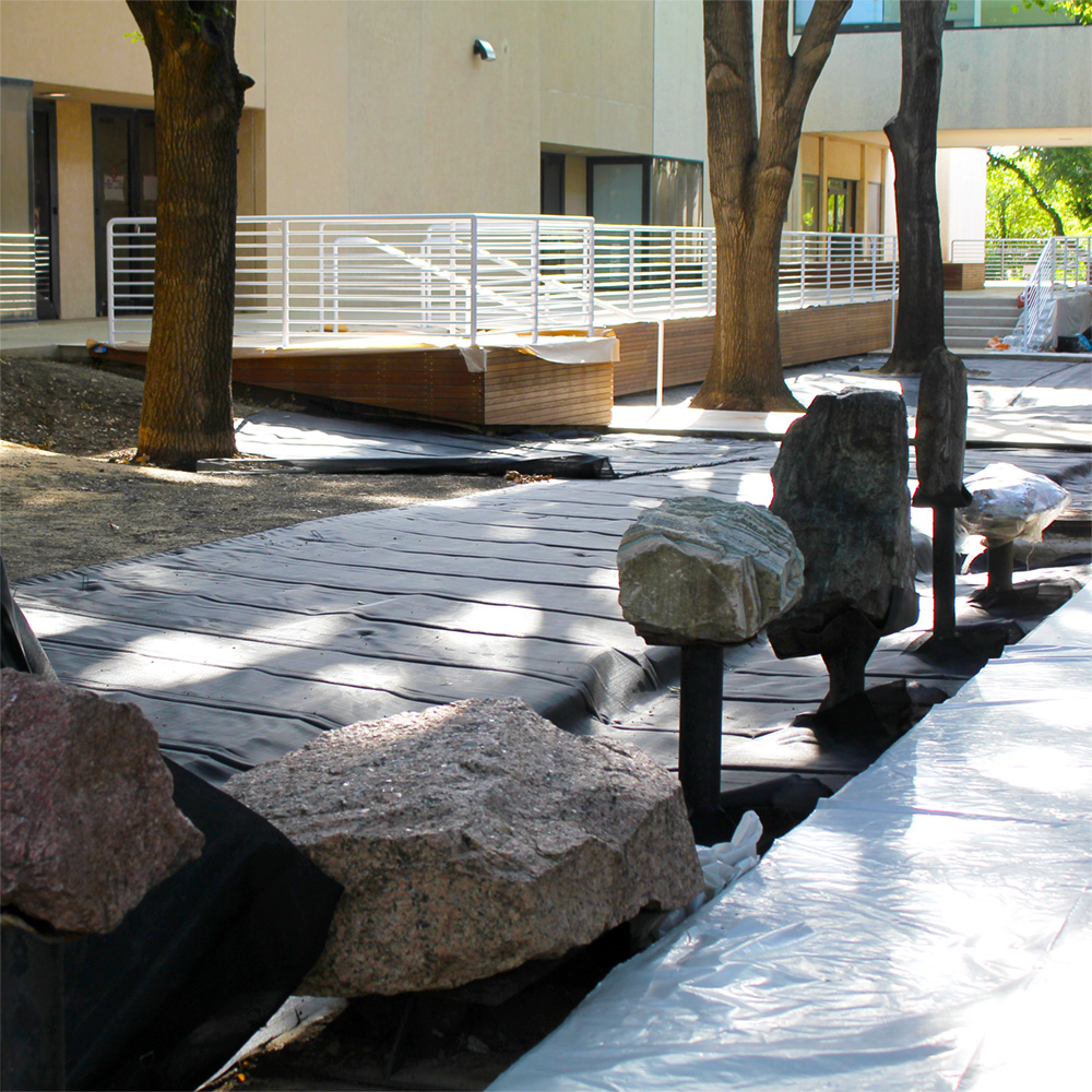 A tree-shaded space between two buildings containing bare earth and several large rocks mounted on iron pillars.