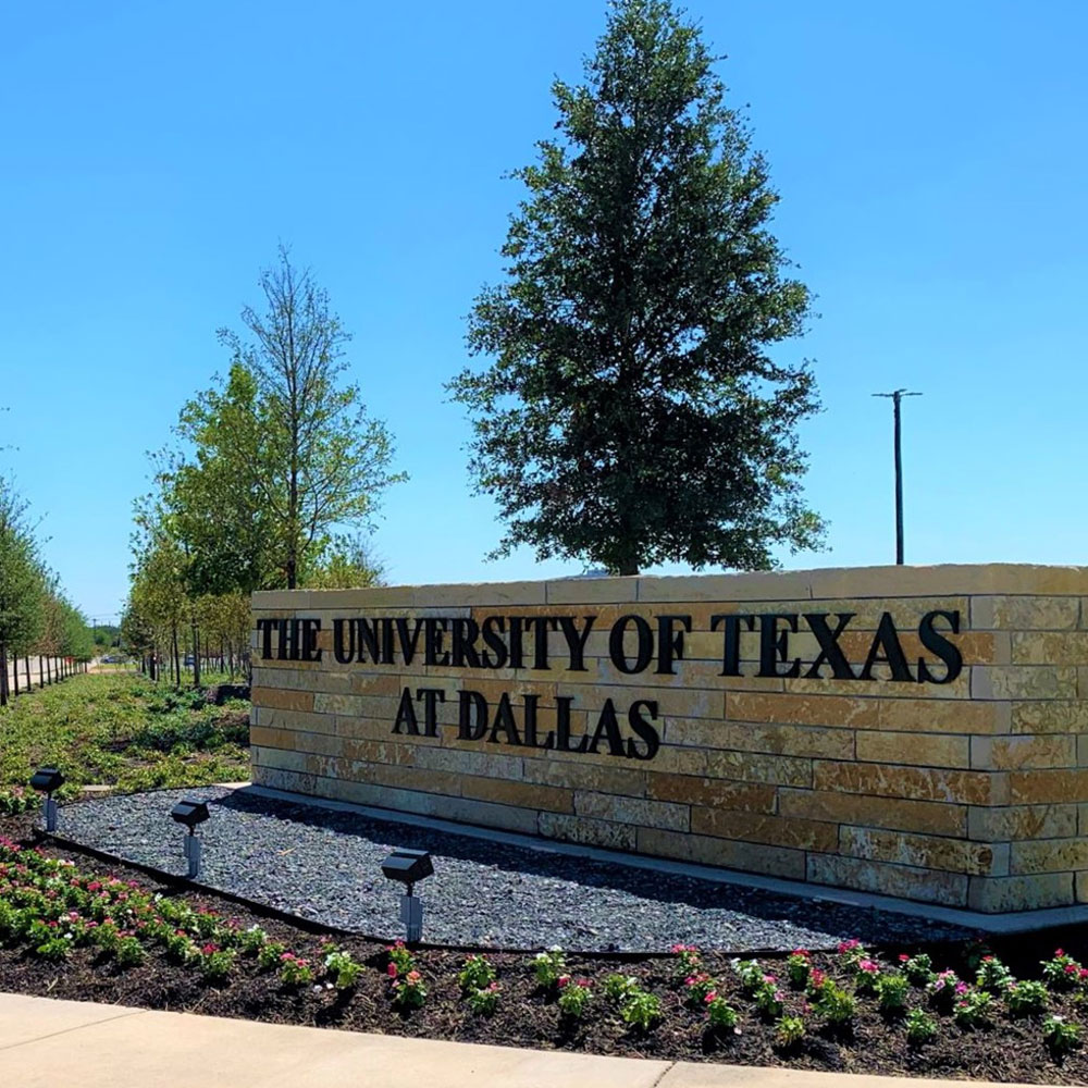 At a tree-lined street corner in the middle of a flower bed stands a large half-height wall of pale stones, on which are mounted metal letters which read “The University of Texas at Dallas”.