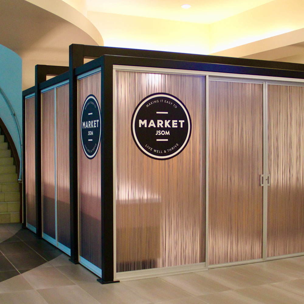 A room-sized box constructed from dark metal beams and translucent pink panels, illuminated from within, bearing a sign which reads “Market - JSOM”.