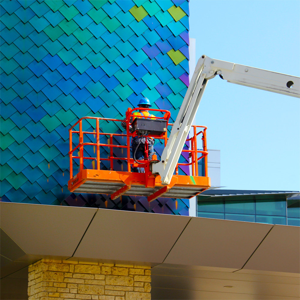 A worker with a safety vest and hardhat rides in the basket of a cherry-picker in front of a wall of overlapping diamond-shaped metal scales.