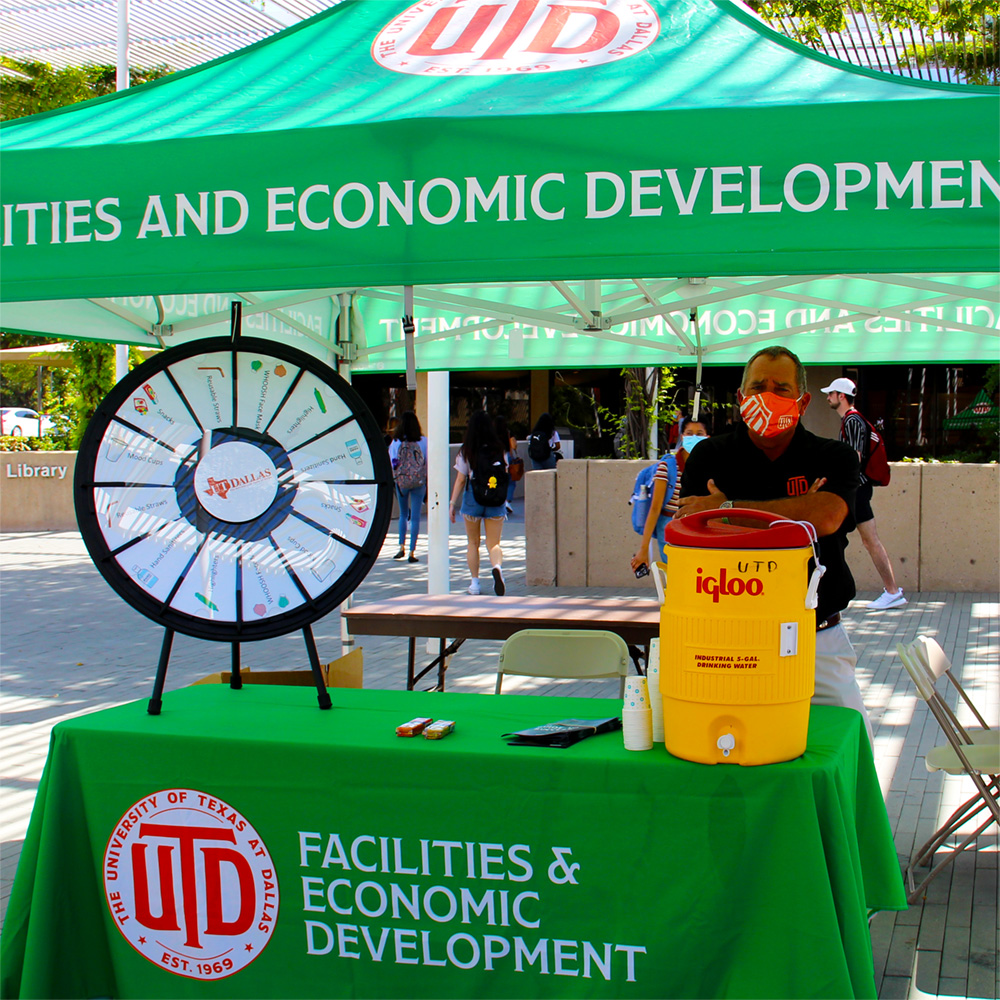 A man in a COVID face mask stands under a tent at a table with a water cooler, a stack of maps and game wheel.