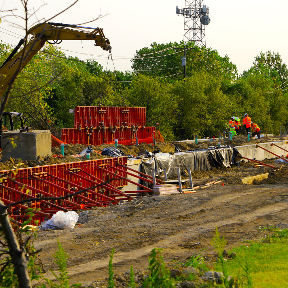 A crane positions concrete molds around the growing foundation of an elevated concrete platform, set in the middle of a dirt track carved out of a wooded area.