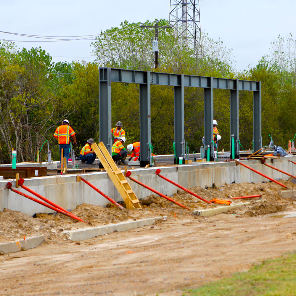 Workers inspect a metal framework coming up from the growing foundation of an elevated concrete platform, set in the middle of a dirt track carved out of a wooded area.