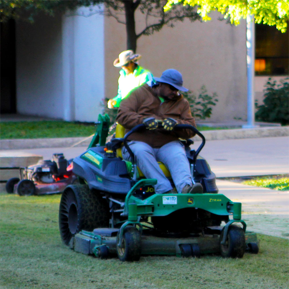 Two groundskeepers, one on a riding mower and another with a push mower, mow their way down a courtyard lawn.