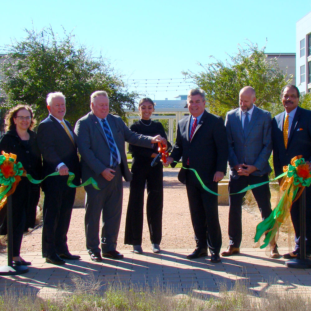 Two people cut a suspended ribbon using a giant pair of scissors as four others look on.