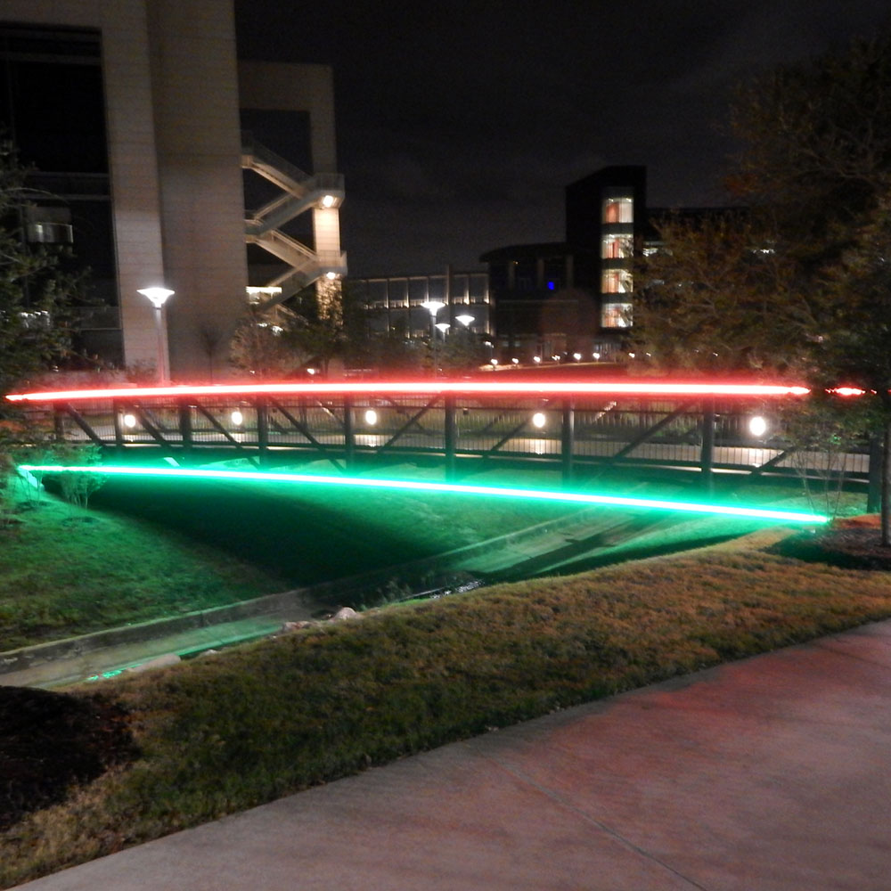 Orange LED lights run along the top rail of a black bridge spanning a creek, while a row of green lights run along the bottom.