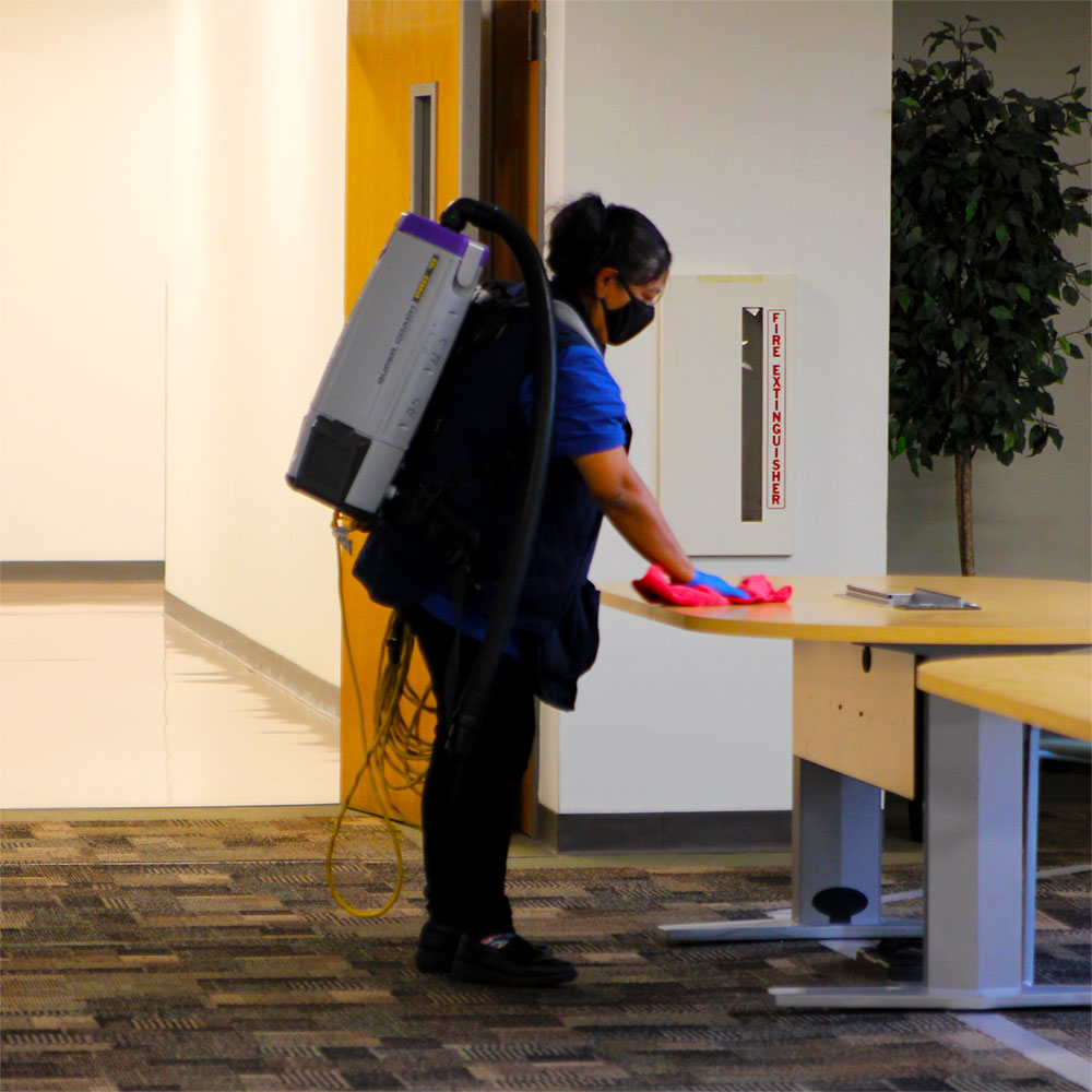 A woman in a facemask and gloves with a vacuum cleaner strapped to her back wipes down tables and chairs.