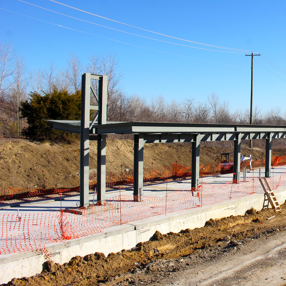 Metal girders forming pillars and a roof above a concrete platform in the middle of a a dirt track surrounded by trees.