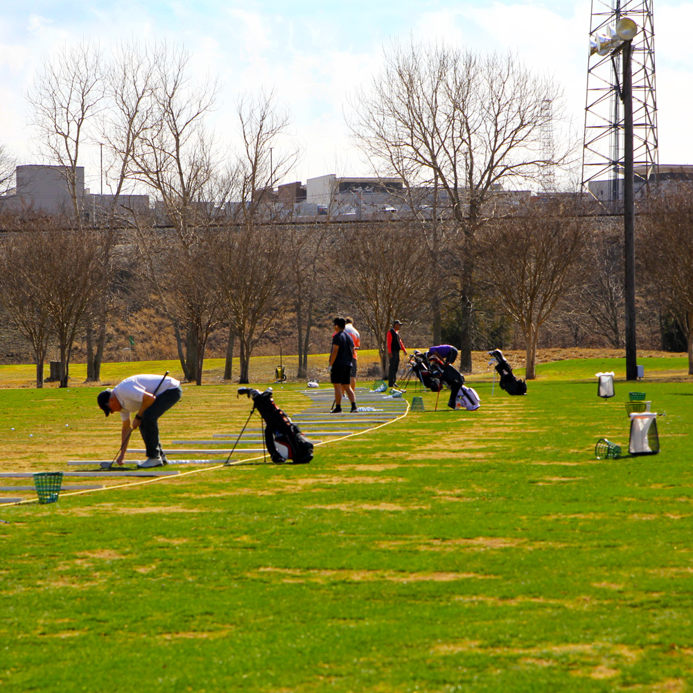 A group of golfers with their bags of clubs line up along a practice range. The tops of UT Dallas buildings can be seen in the background behind a hill.