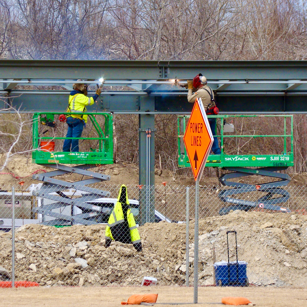 A pair of workers in welding gear atop small cherry-pickers weld beams onto the developing roof structure of what will become a train station.