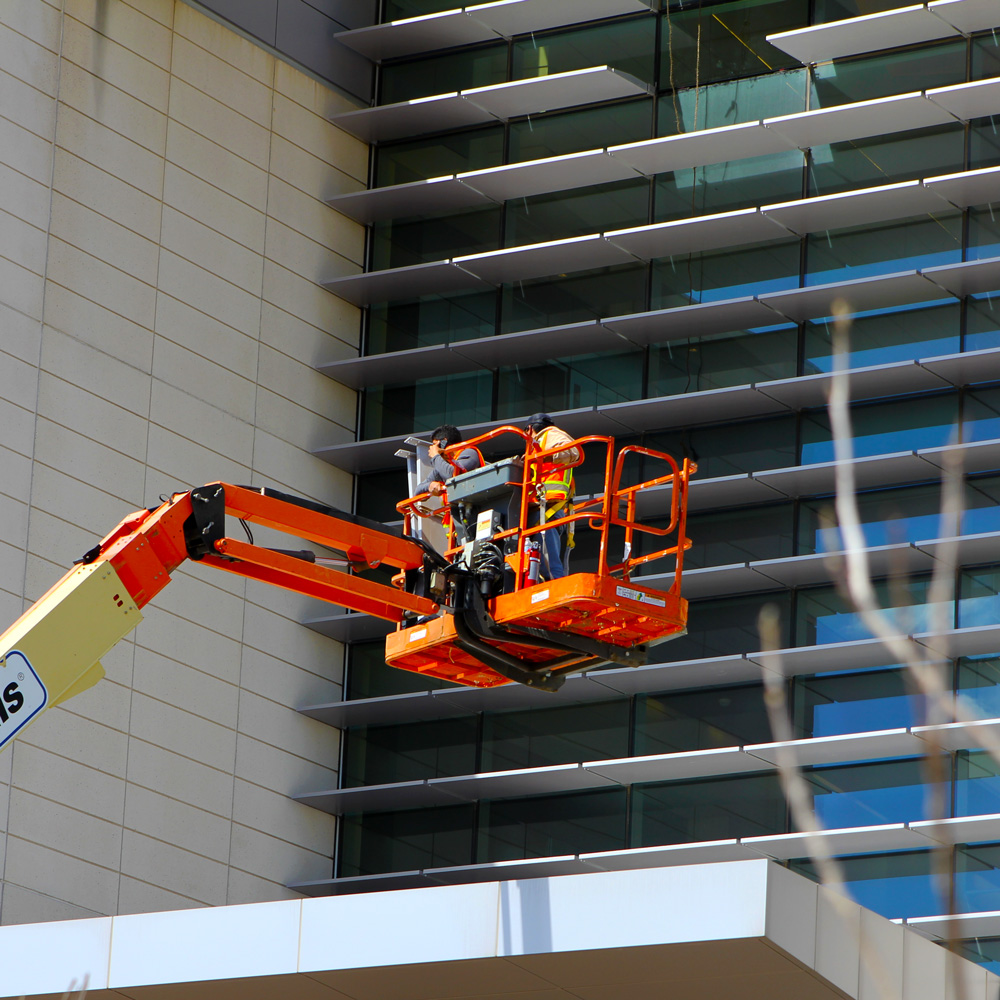 Two workers ride a chery-picker up the side of a building that has a missing panel, bringing a replacement panel along with them.