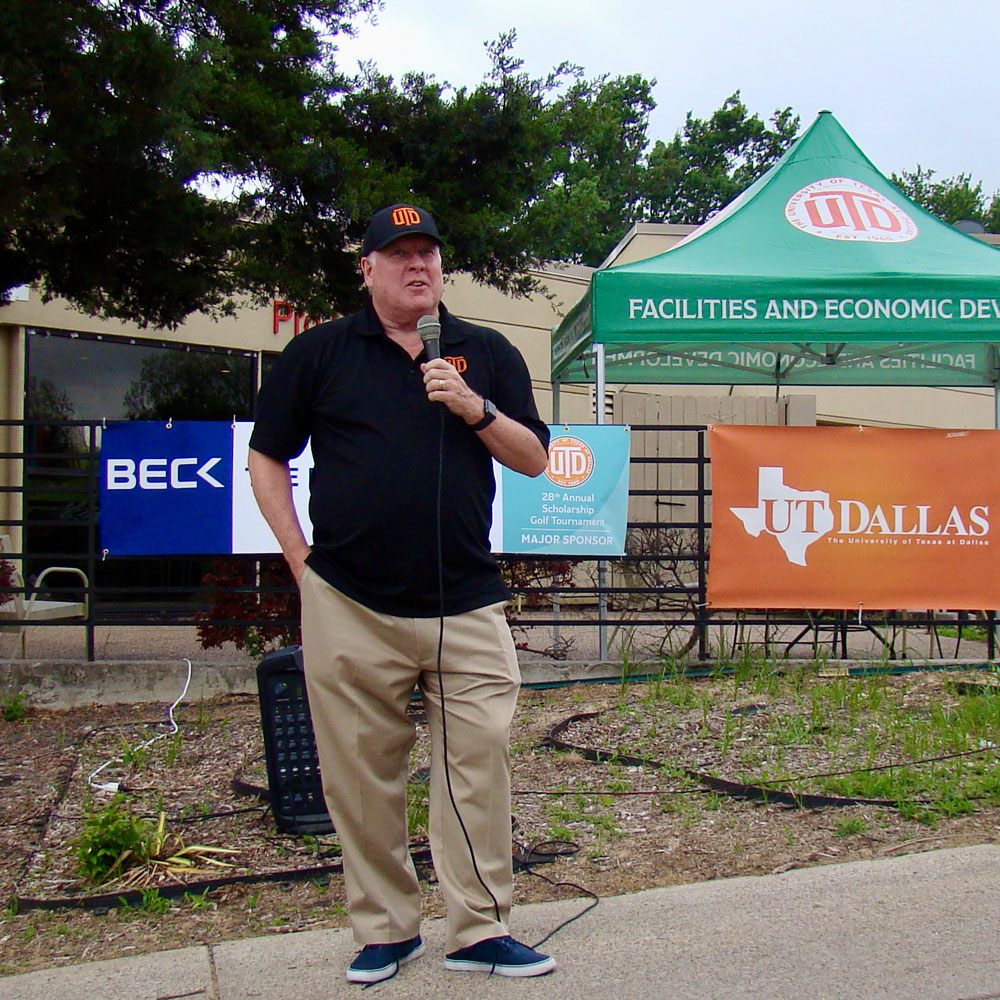 Dr. Richard C. Benson, in a UT Dallas hat and shirt, speaks in front a variety of banners for the UT Dallas Scholarship Golf Tournament.