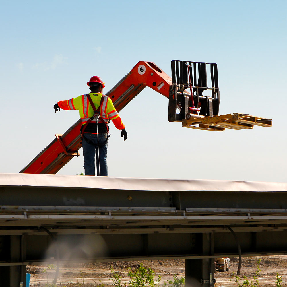 A construction worker stands atop a roof being added to the metal skeleton of a structure, directing the actions of a forklift that has raised a pallette to the level of the new roof.