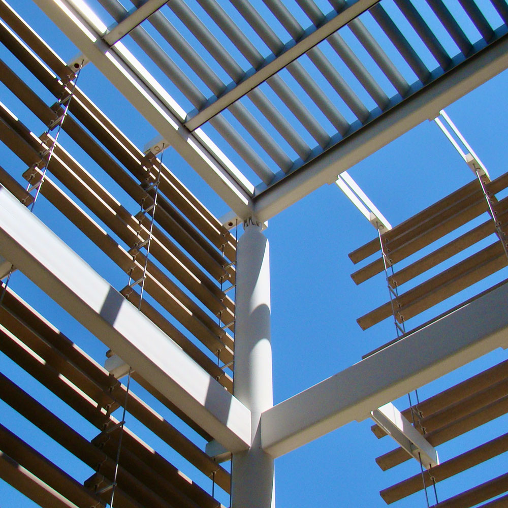 A series of horizontal strips suspended from a white metal frame outside of a building, meeting at a corner, as seen from below, with light filtering in through the spaces between the strips.