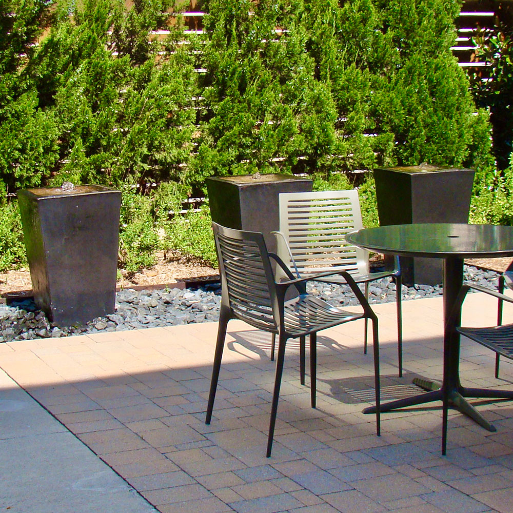 The corner of a garden walled with horizontal wooden slats, bordered by greenery, and paved in reddish tiles. Along one wall, three modern minimalist metal fountains bubble water down their sides. In the foreground are dark metal tables and chairs.