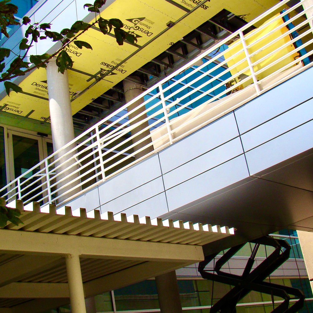 An outdoor walkway between two buildings, clad in metal and supported by concrete pillars, seen from an angle. Some material from its roof has been pulled back to show insulating materials underneath. From below, a cherry-picker extends up to its level.