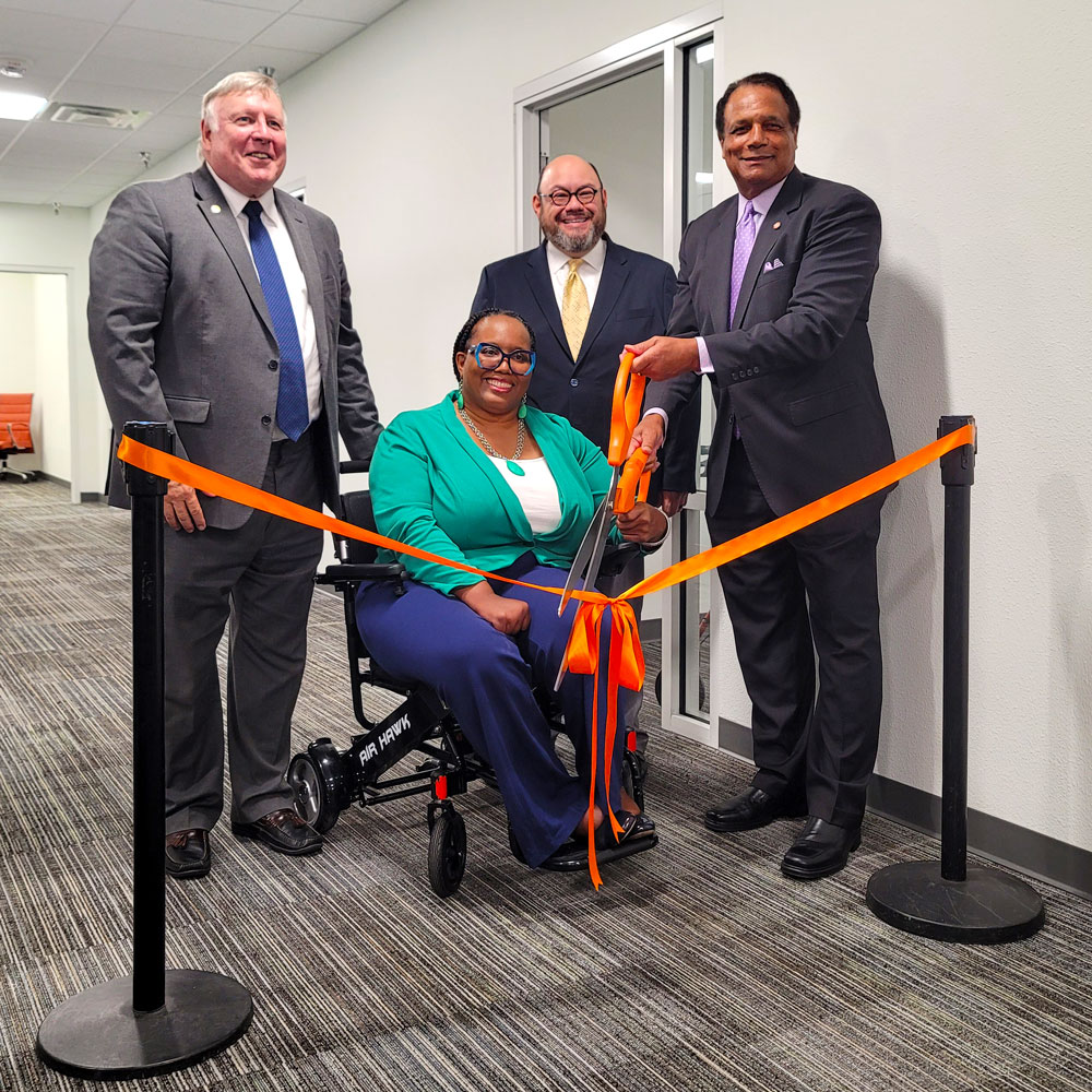 Four people in an office lobby prepare to cut a ribbon suspended in front of them using a giant pair of scissors.