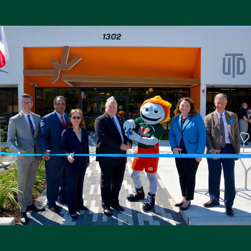 A group of people and the University mascot, an anthropomorphic comet, stand in front of a building as they prepare to cut a blue ribbon suspended in front of them.