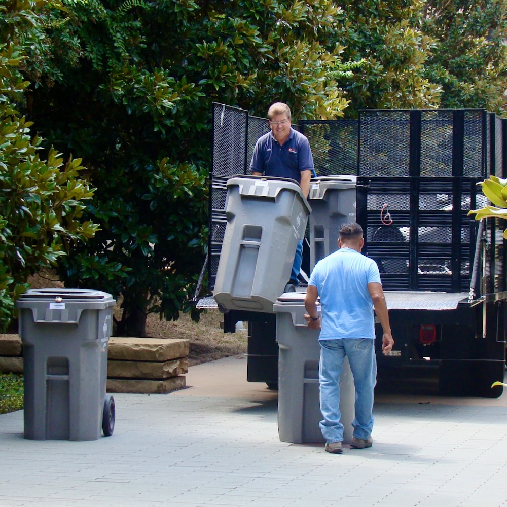 Two workers loading large trash bins into the back of a truck.