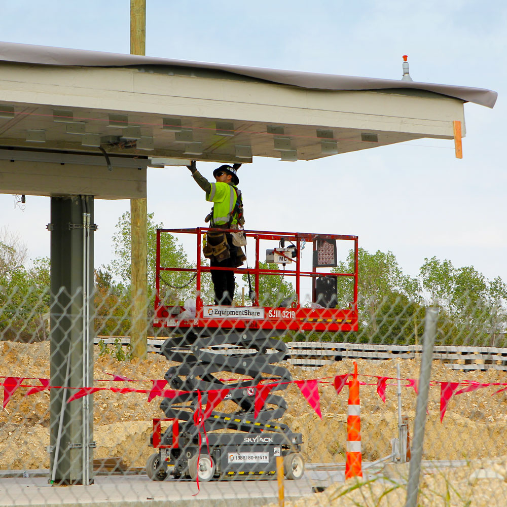 A construction worker standing on top of a cherry-picker while working on the underside of a train platform roof.