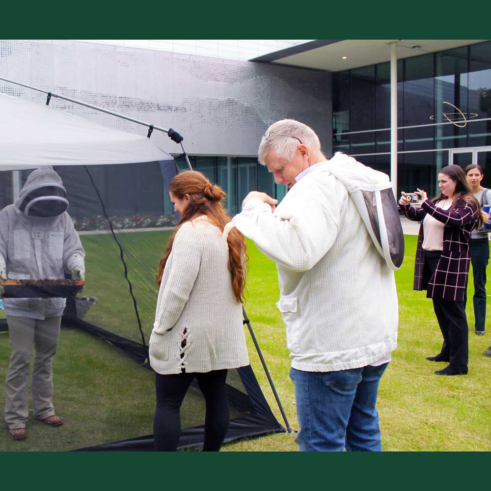 A person in a beekeeper’s protective gear holds out a honeycomb full of bees while standing within a net tent. A crowd safely outside the tent is captivated by the presentation. Among them, another person is donning beekeeper gear in preparation to enter the tent.