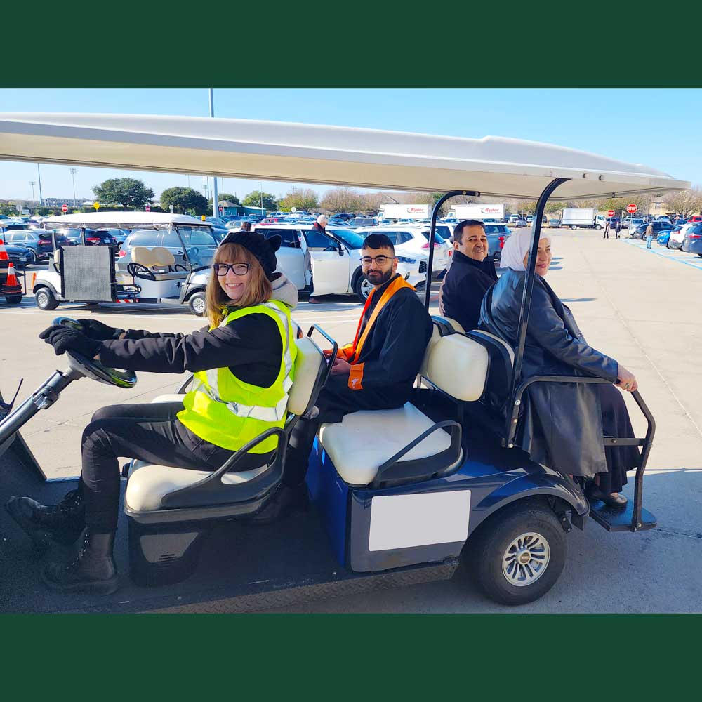 A UT Dallas golf cart style shuttle pulls out of a parking lot, bearing a UTD driver, a student in a graduation gown, and two of his family members.