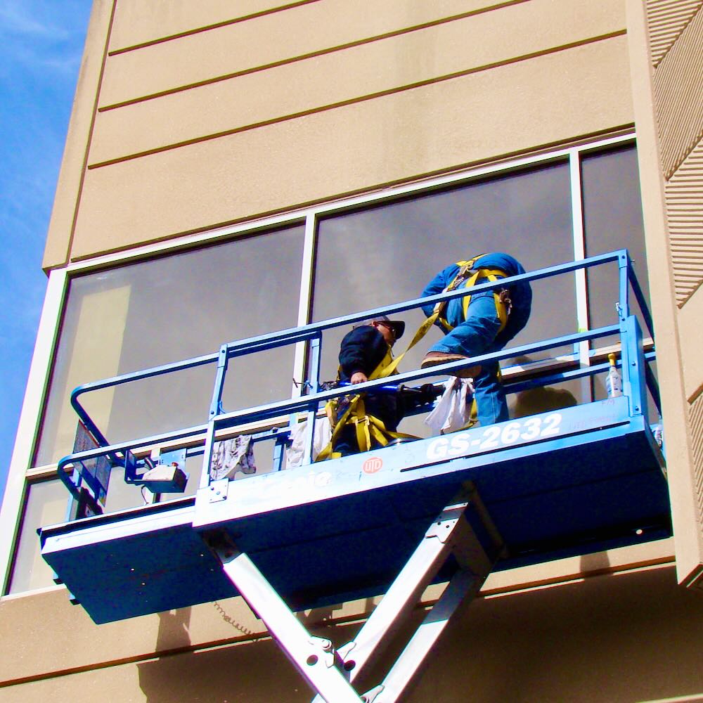 Two workers atop a cherry-picker repair the seal on a glass window set high upon a textured concrete wall.