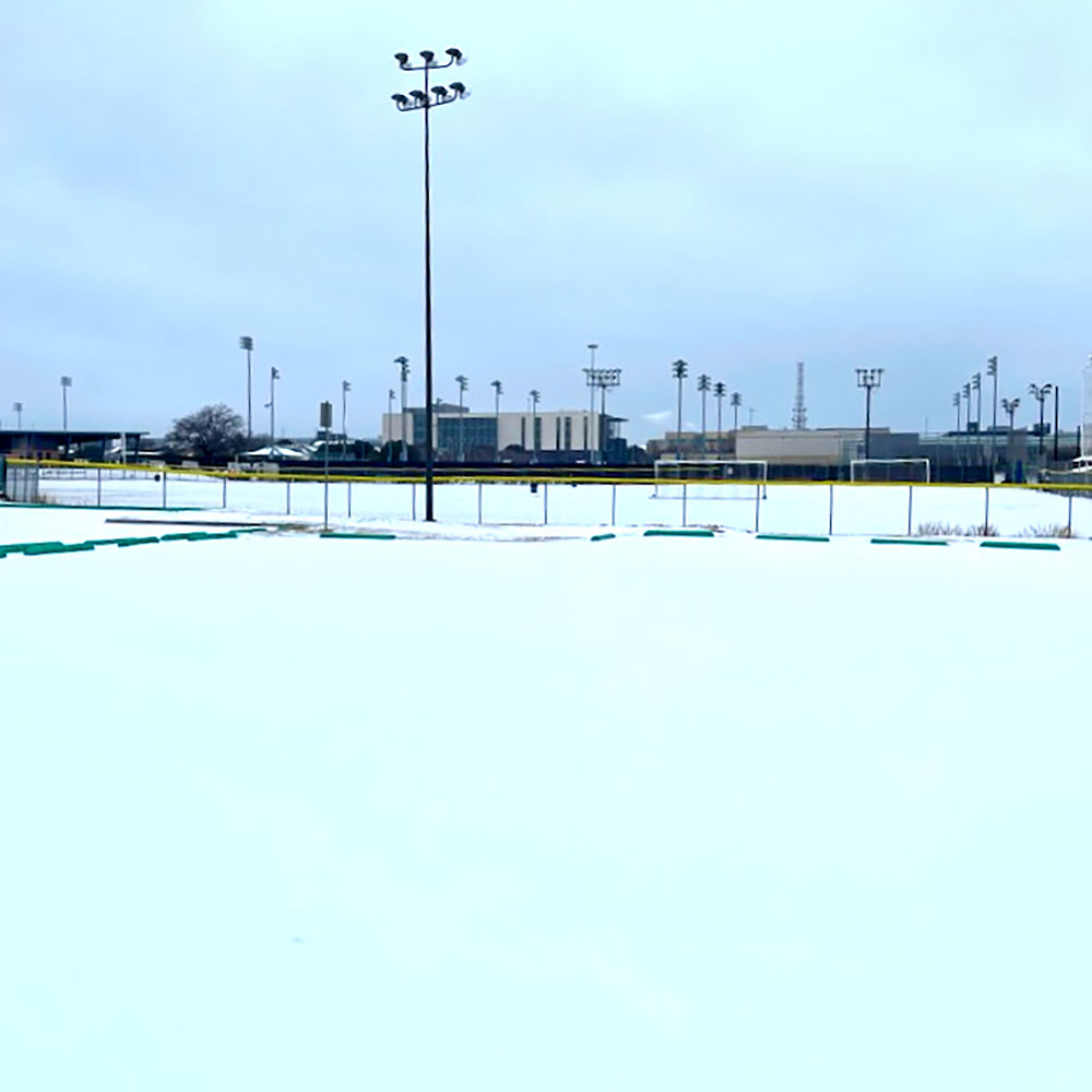 A modern steel and stone building rises above several low concrete buildings on the horizon below an overcasat sky. In the foreground are the fences surrounding athletic fields, but they, along with every other piece of ground, are buried in ice and snow for as far as the eye can see.