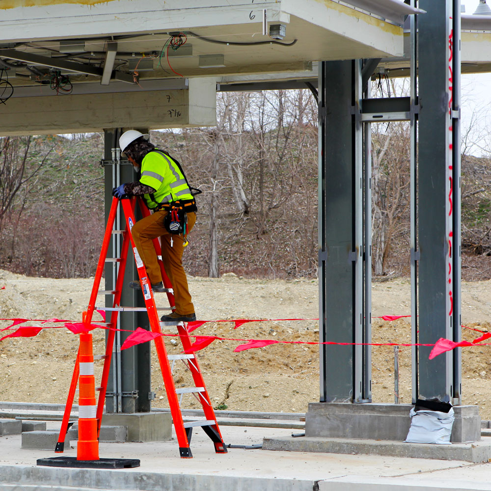 A worker in protective gear climbs a ladder to work on the underside of the roof on a train platform that is under construction.