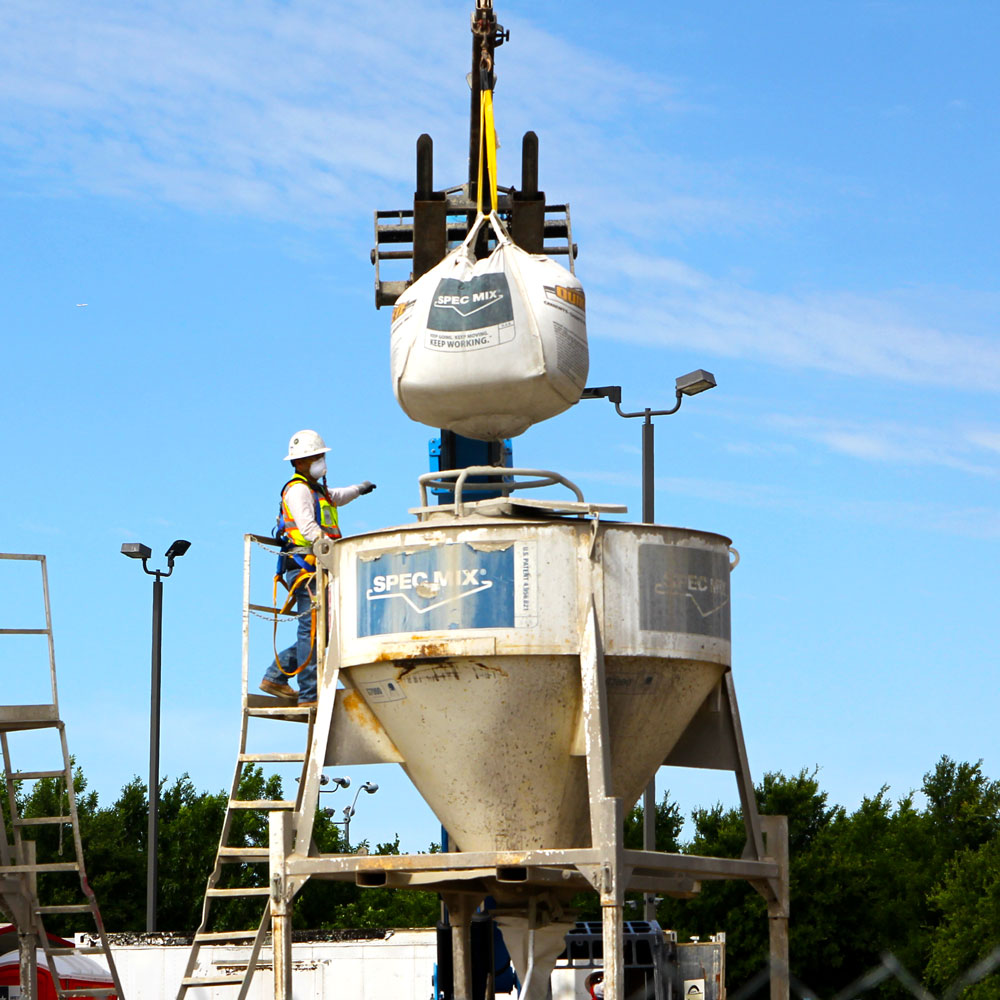 A huge bag of concrete mix is lowered by crane into an upright concrete mixer as a construction worker directs it into position.