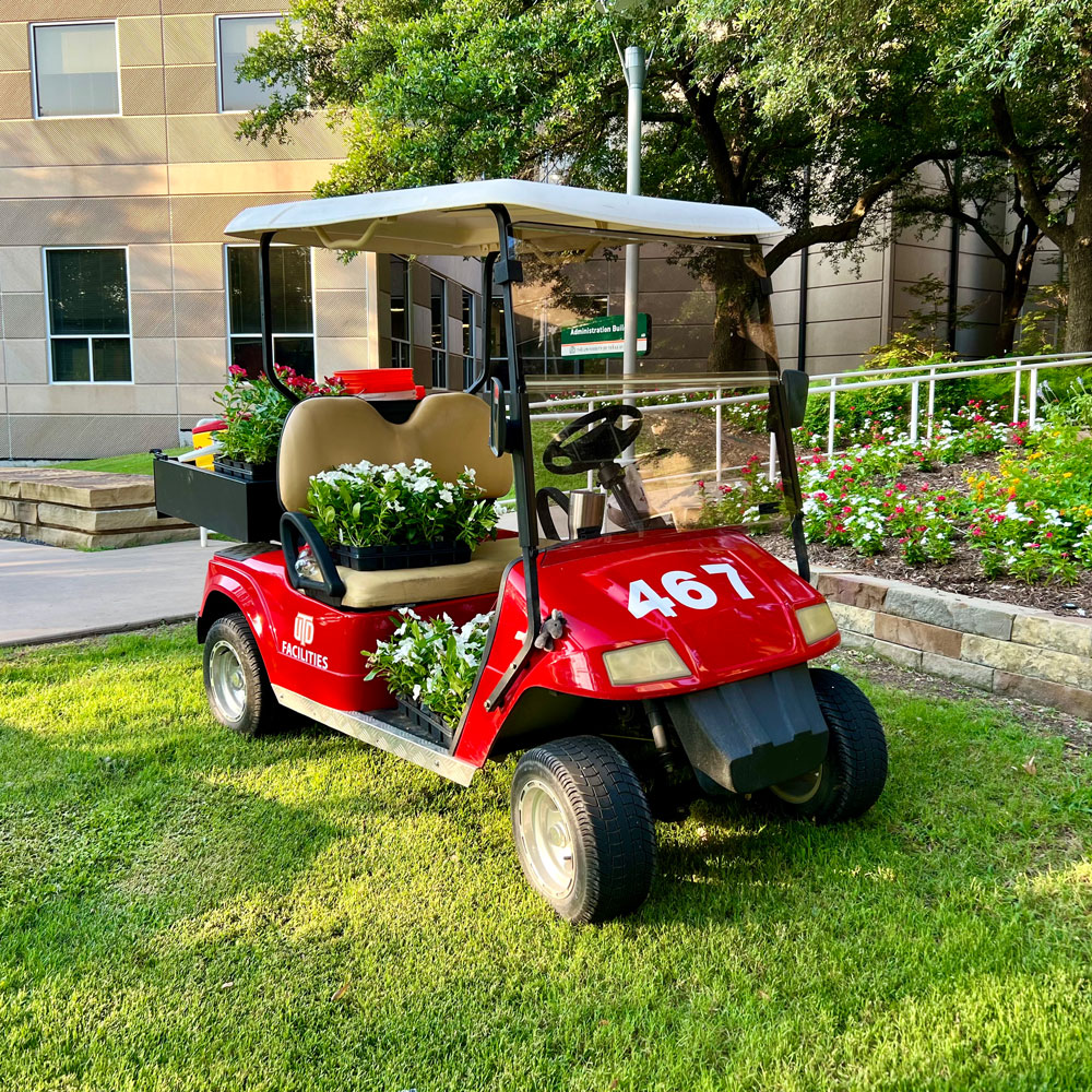 A red cart parked in front of raised flower beds, overflowing with boxes of potted flowers that are ready to be transplanted.
